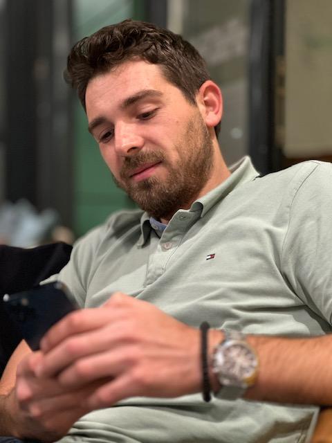 A young man with short curly hair and a beard looks down at his smartphone, wearing a light sage green Tommy Hilfiger polo shirt, a silver watch, and a beaded bracelet, seated in a relaxed indoor setting.