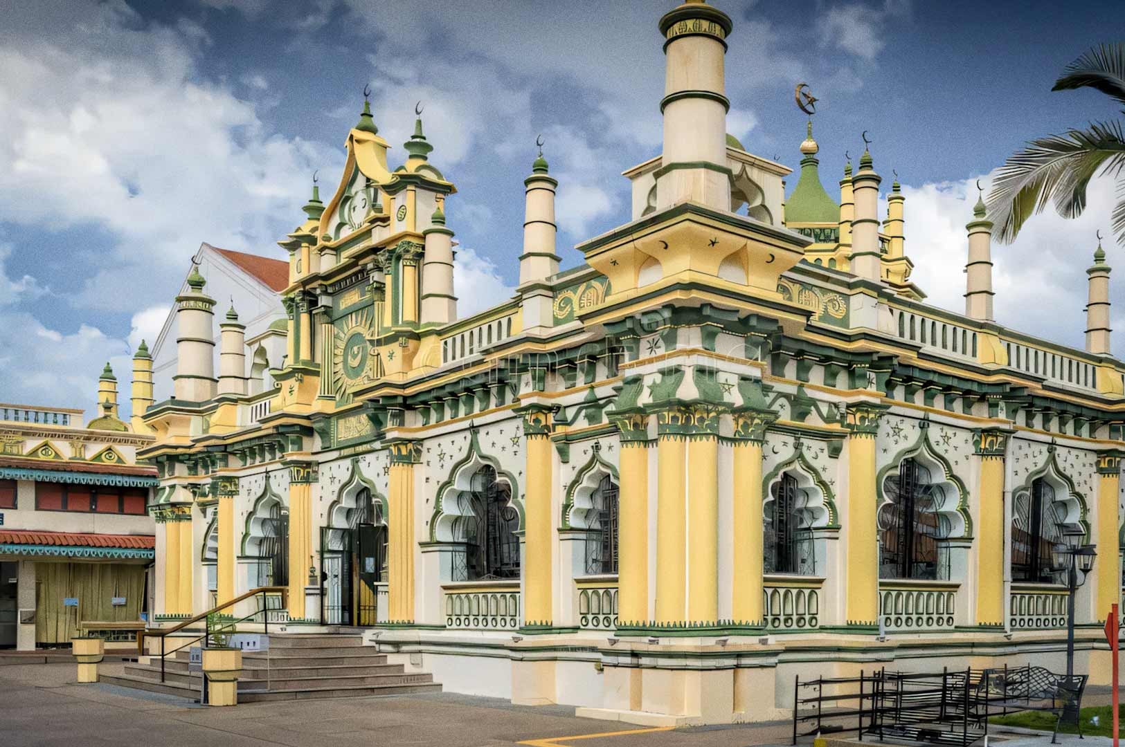 A view of the Abdul Gaffoor Mosque featuring its distinctive yellow-and-green façade, ornate arches, and multiple decorative minarets under a partly cloudy sky.