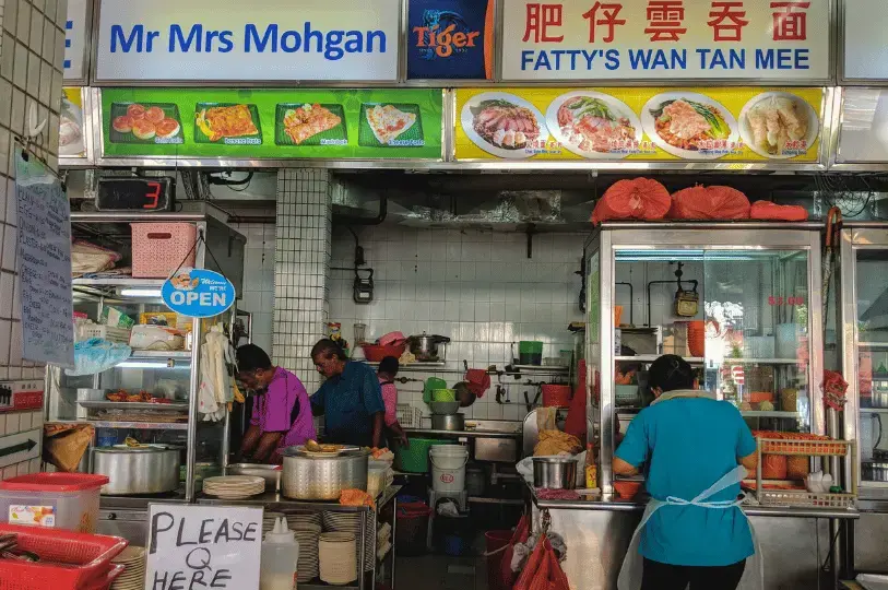 A bustling hawker stall named Mr Mrs Mohgan, showing cooks busy preparing fresh roti prata in the kitchen beneath a brightly lit menu board.