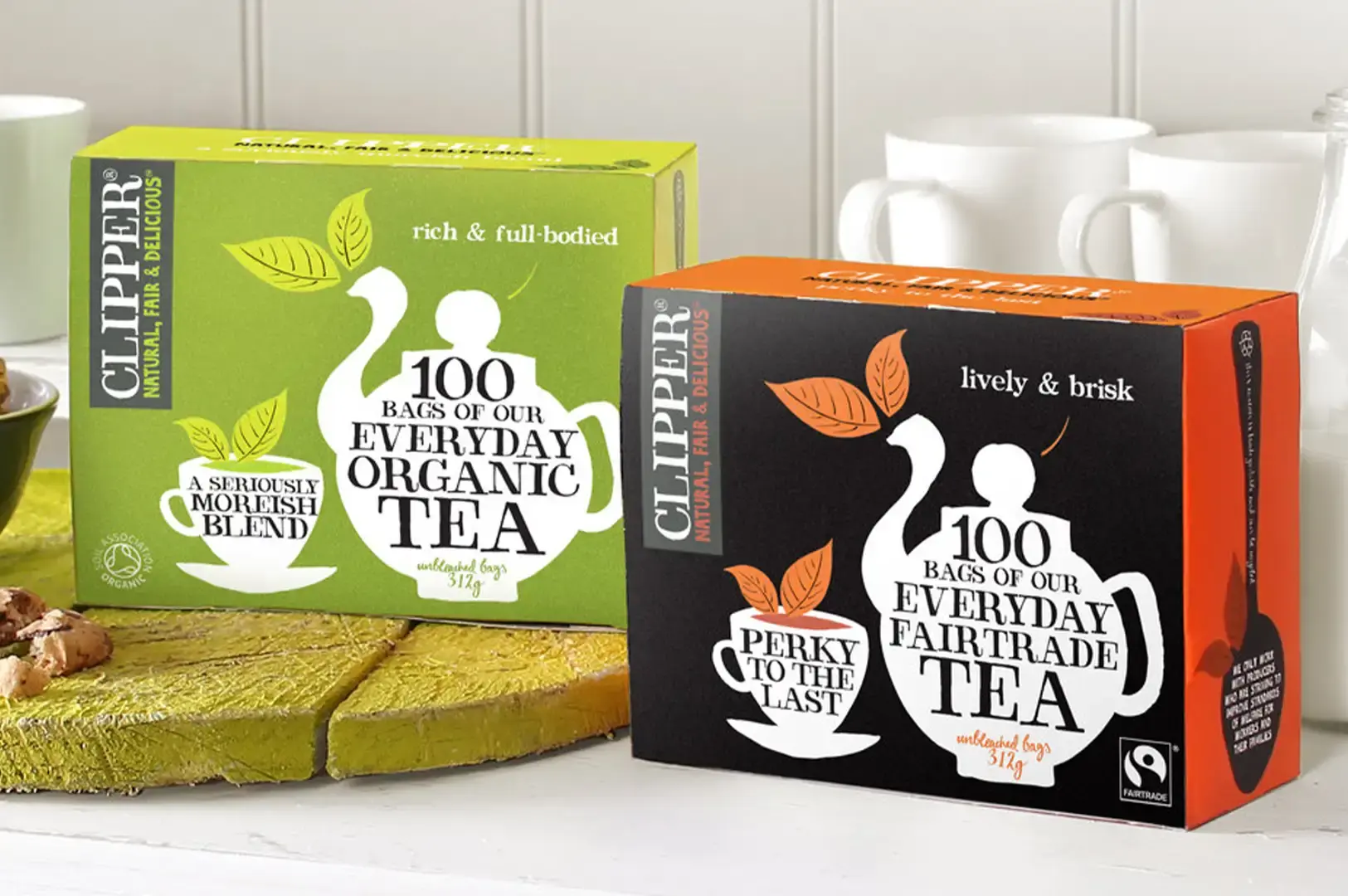 A close-up side-by-side display of two large 100-bag Clipper tea boxes sitting on a white surface next to a textured yellow board. On the left is the green "Everyday Organic Tea" box featuring a green teapot motif, and on the right is the black and orange "Everyday Fairtrade Tea" box featuring a black teapot illustration, with white ceramic mugs subtly blurs in the background.