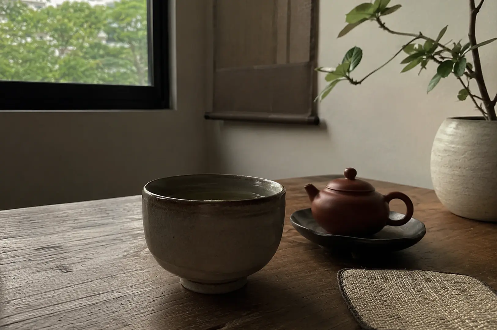 A moody, close-up shot focuses on a small tea arrangement on a weathered wooden table. A large, thick-walled ceramic cup with a rough, beige-and-grey glaze sits in the foreground. Behind it and to the right, a small, smooth reddish-brown clay teapot rests on a tiny matching saucer. Further right, a round, matte white ceramic vase holds a branch with sparse green leaves. In the upper left background, a dark-framed window reveals green trees outside, while a simple brown fabric scroll hangs on the plain wall beside it, enhancing the minimalist atmosphere.
