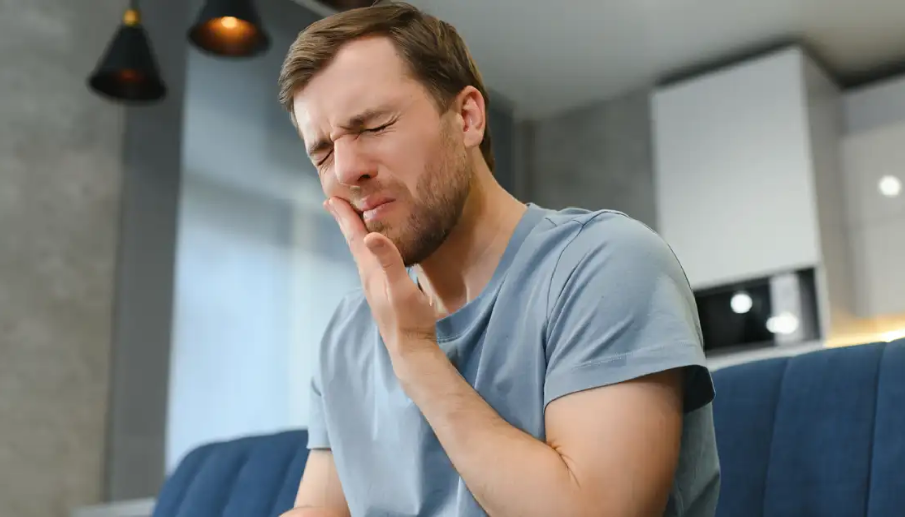 Mann med skjegg holder seg til kinnet og viser tydelige tegn på tannverk mens han sitter hjemme i stua.