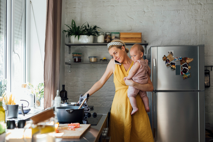 Young mom in a yellow dress holding her baby while cooking and talk on her cell.
