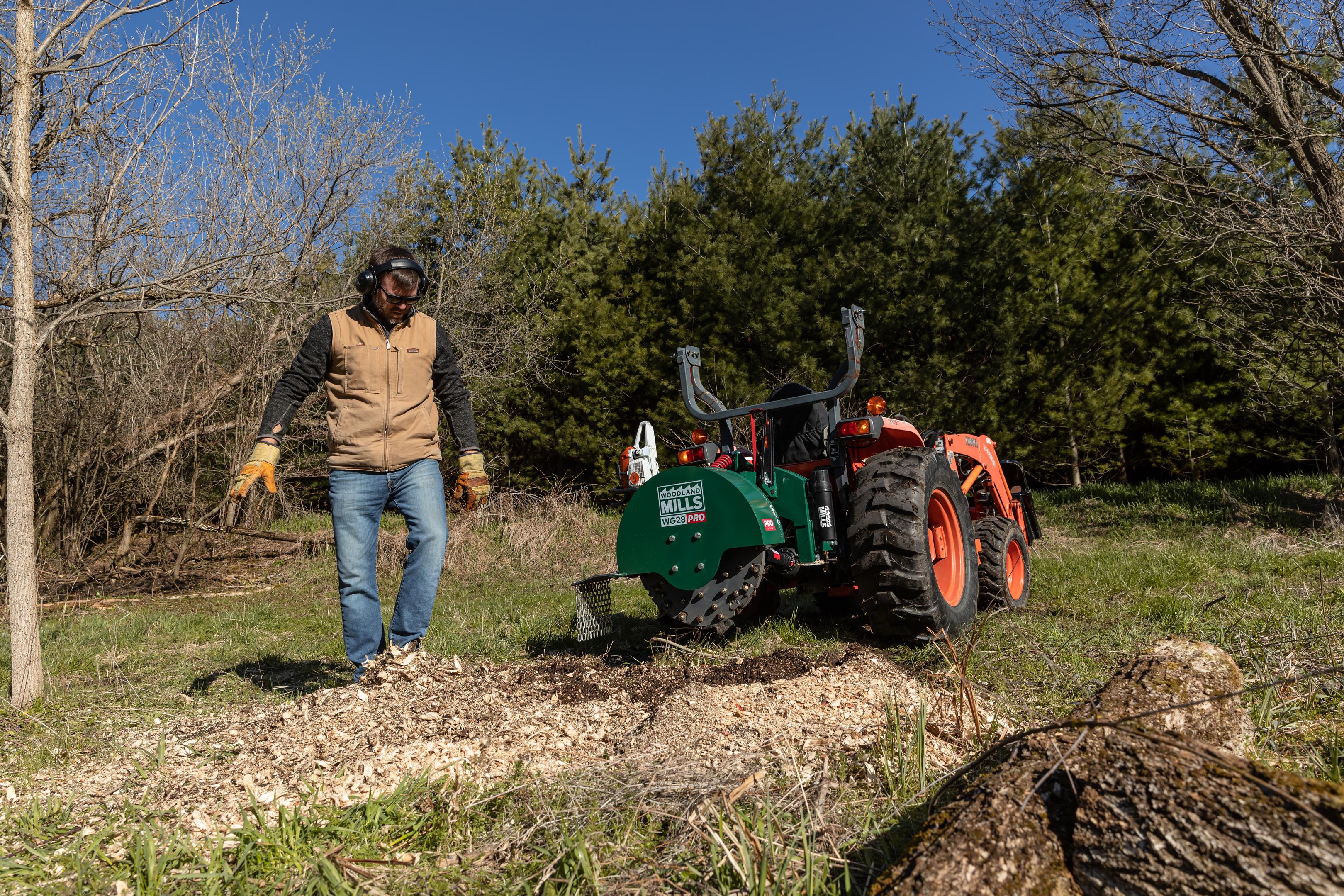 A man standing beside a Woodland Mills WG28 PRO Stump Grinder on the back of a tractor.