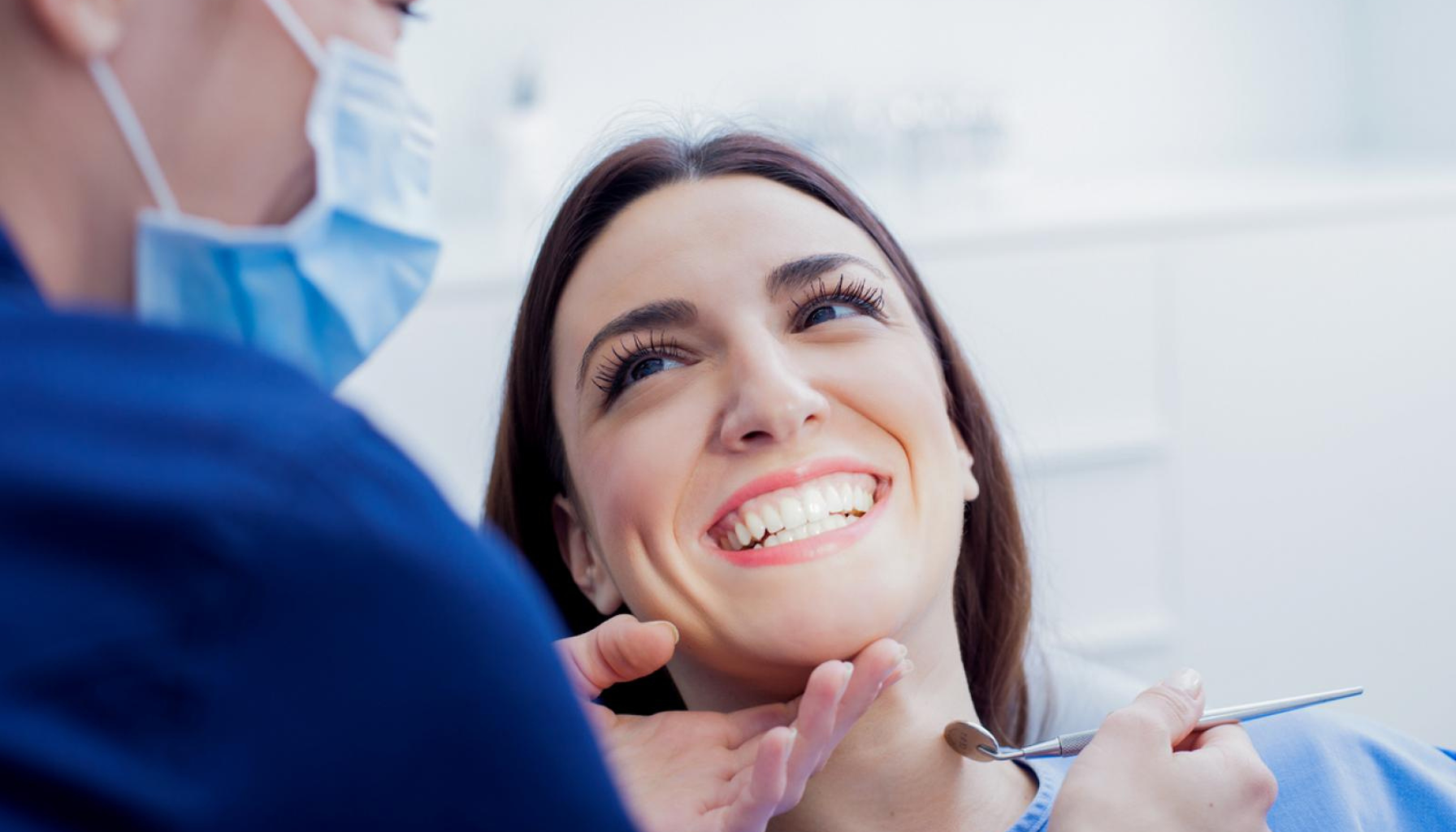 Kvinne smiler bredt mens tannlege med maske undersøker tennene hennes og holder en tannlegeinstrument i munnen hennes. 