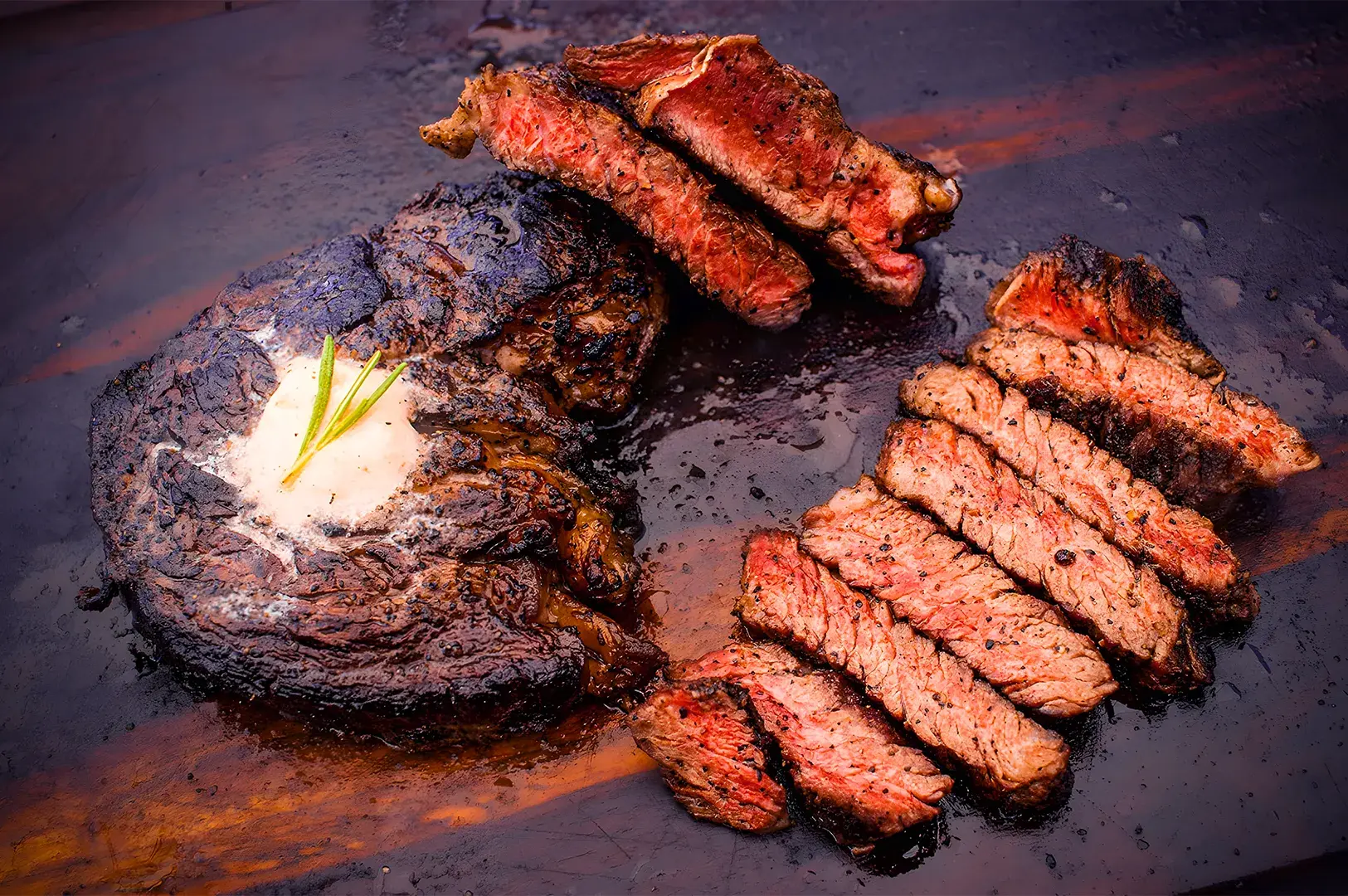 Grilled ribeye steak on a dark wooden board, topped with melted butter and herbs, alongside juicy, sliced pieces, highlighting its charred and tender texture.