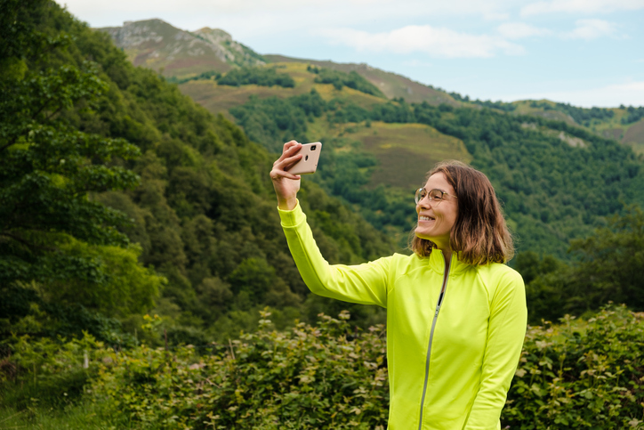 Happy dark-haired young woman in a lime-colored jacket snapping a selfie on a hiking trail. 