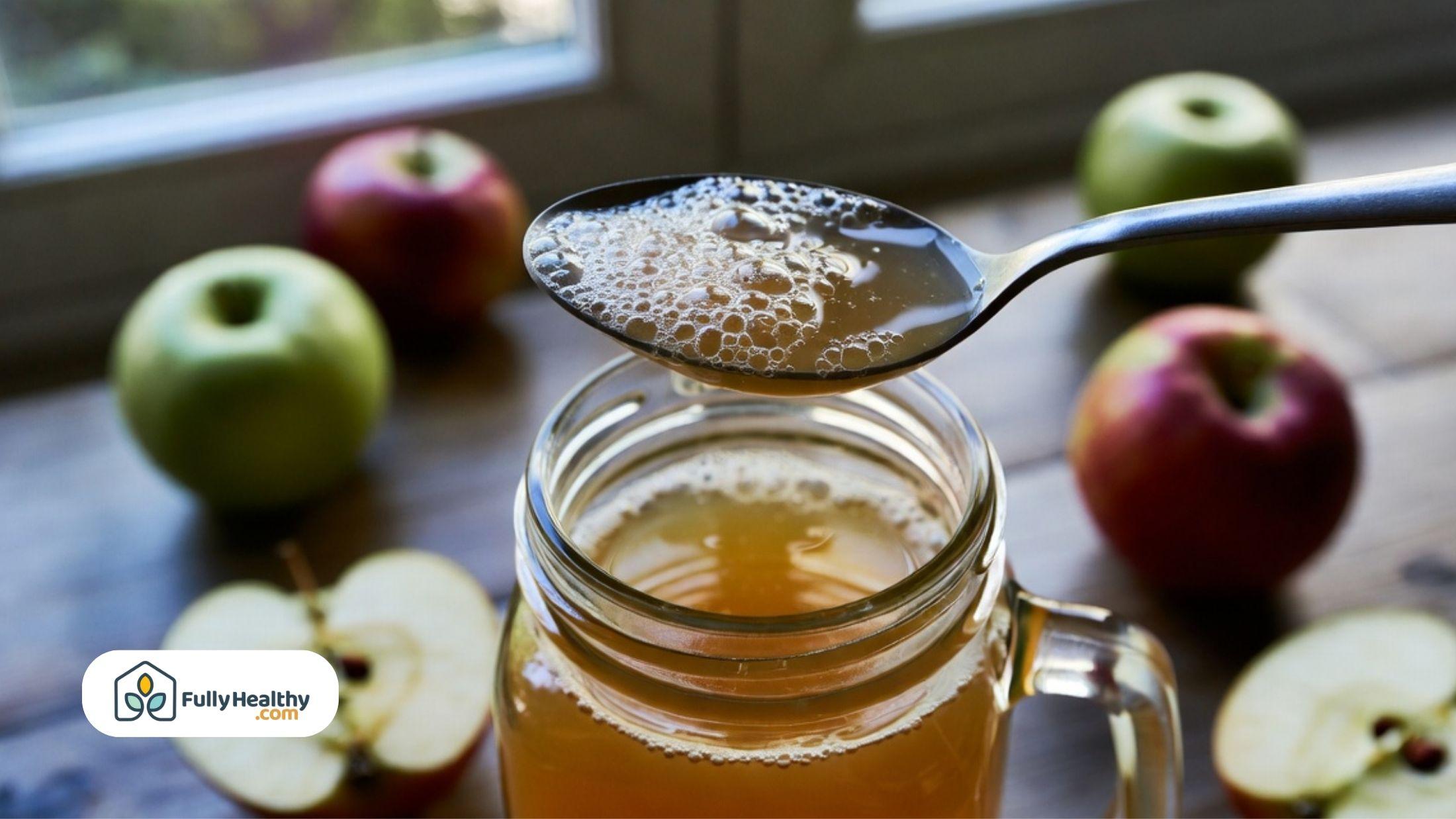 Spoon lifting apple cider from a glass jar with fresh apples on a wooden table.