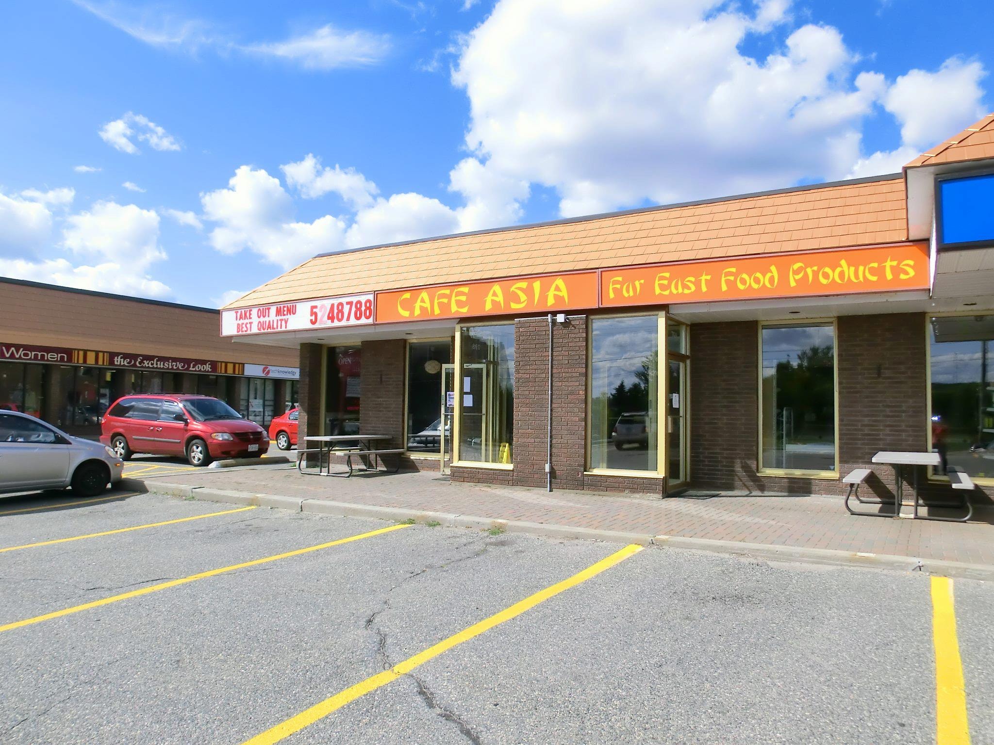 A strip mall with a storefront sign reading “Cafe Asia Far East Food Products” and parked cars in front under a partly cloudy sky, featuring commercial buildings topped with metal roofing.