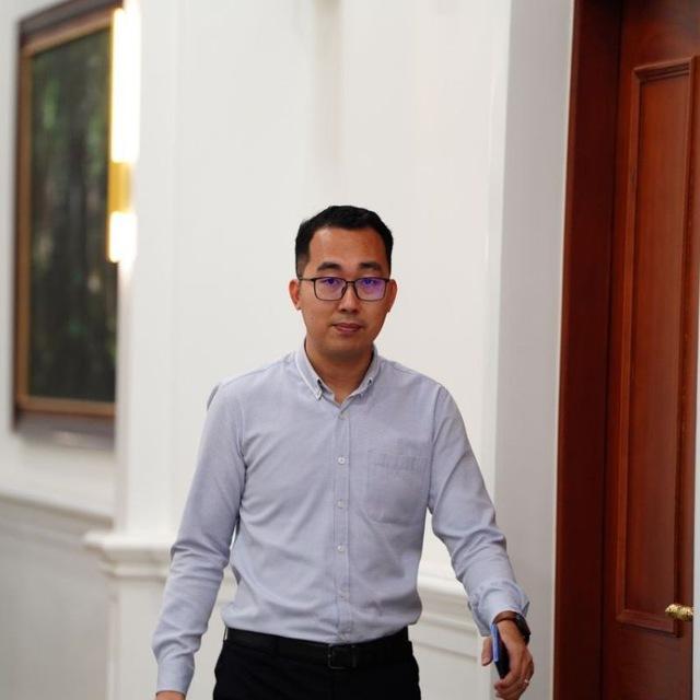 A man wearing rectangular-framed glasses and a light blue button-down shirt walks purposefully through an elegant corridor with white walls, warm sconce lighting, and a dark wood doorframe — conveying a sense of professionalism and quiet confidence in a formal institutional setting.