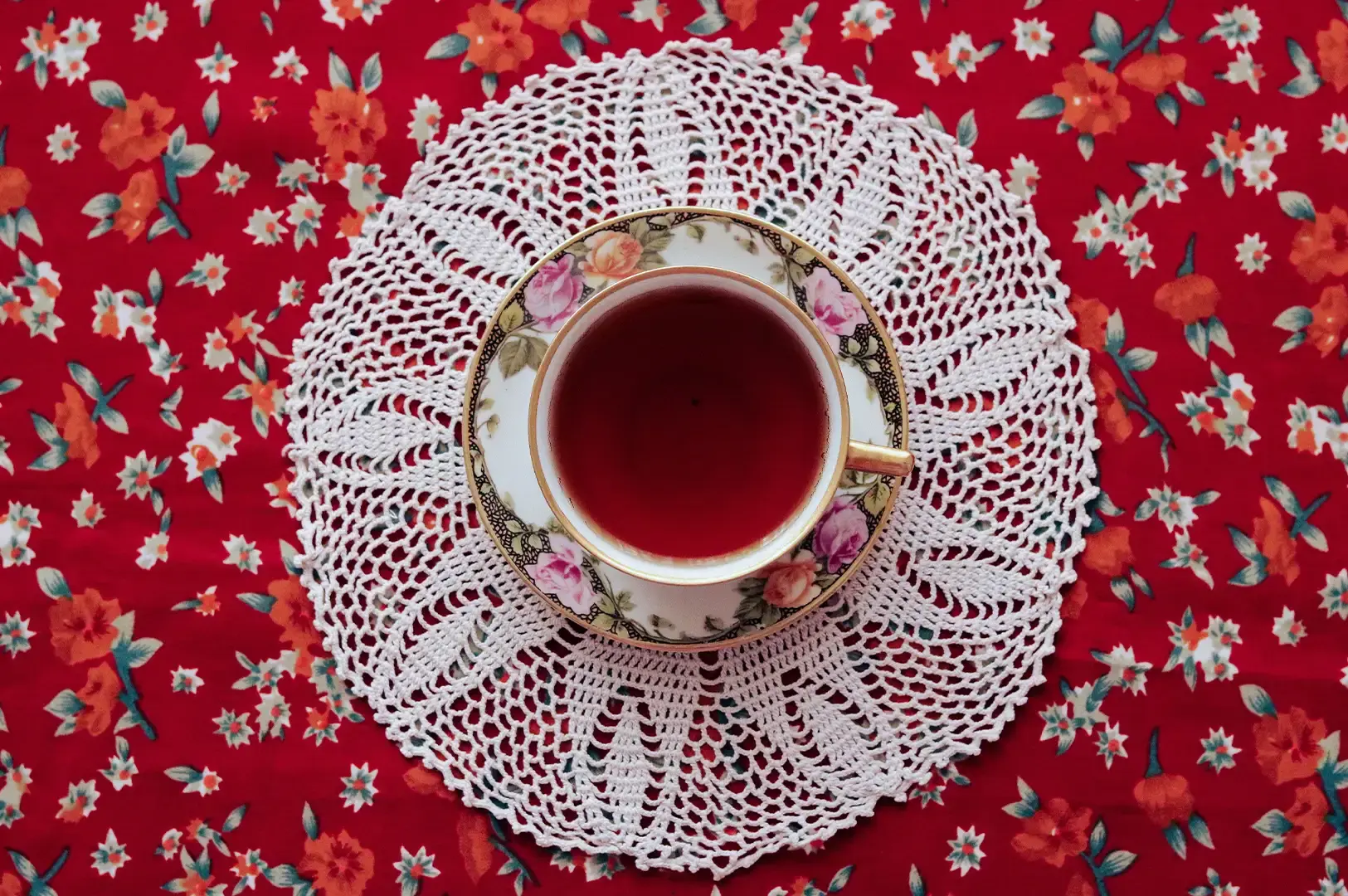 A top-down view shows a gold-trimmed floral teacup filled with dark red tea resting on a white lace doily, set atop a vibrant red tablecloth printed with small orange and white flowers.