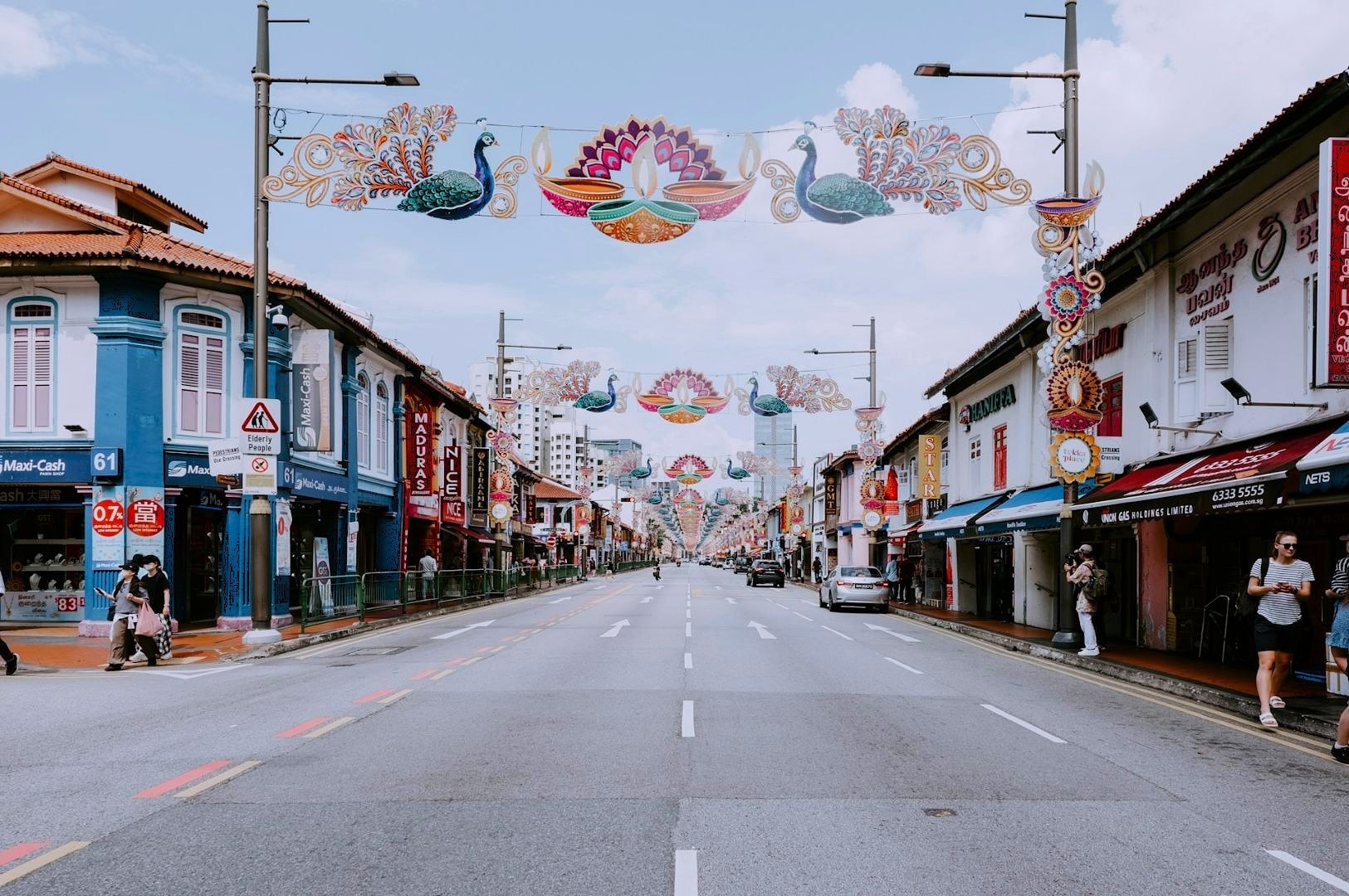 A vibrant street scene with colorful peacock-themed decorations hanging above. Lined with shops, the road is lively yet uncrowded, evoking a festive atmosphere.