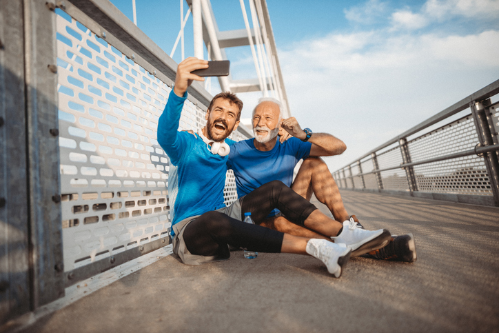Adult son and his dad sitting on the walkway of a bridge after their run and taking a selfie. 