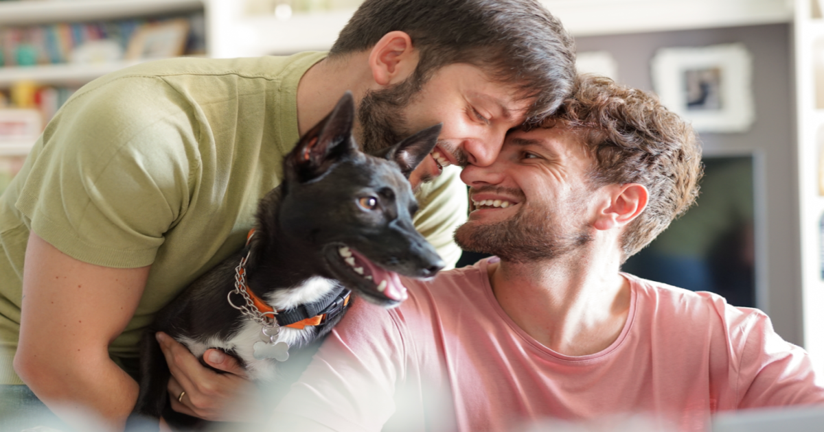 A photograph of a happy gay couple sitting on a park bench with their dog between them, smiling and looking content. The couple appears to be enjoying the outdoors in New York City, possibly after having successfully undergone Gottman Method Couples Therapy. The image conveys the idea of a couple who have improved their emotional connection and relationship through therapy, as evidenced by their happy and content expressions. The presence of their dog suggests a sense of family and companionship. The image suggests that seeking the services of a certified Gottman Method Couples Therapist in New York City can help couples of all kinds improve their relationships and live happier, more fulfilling lives together.