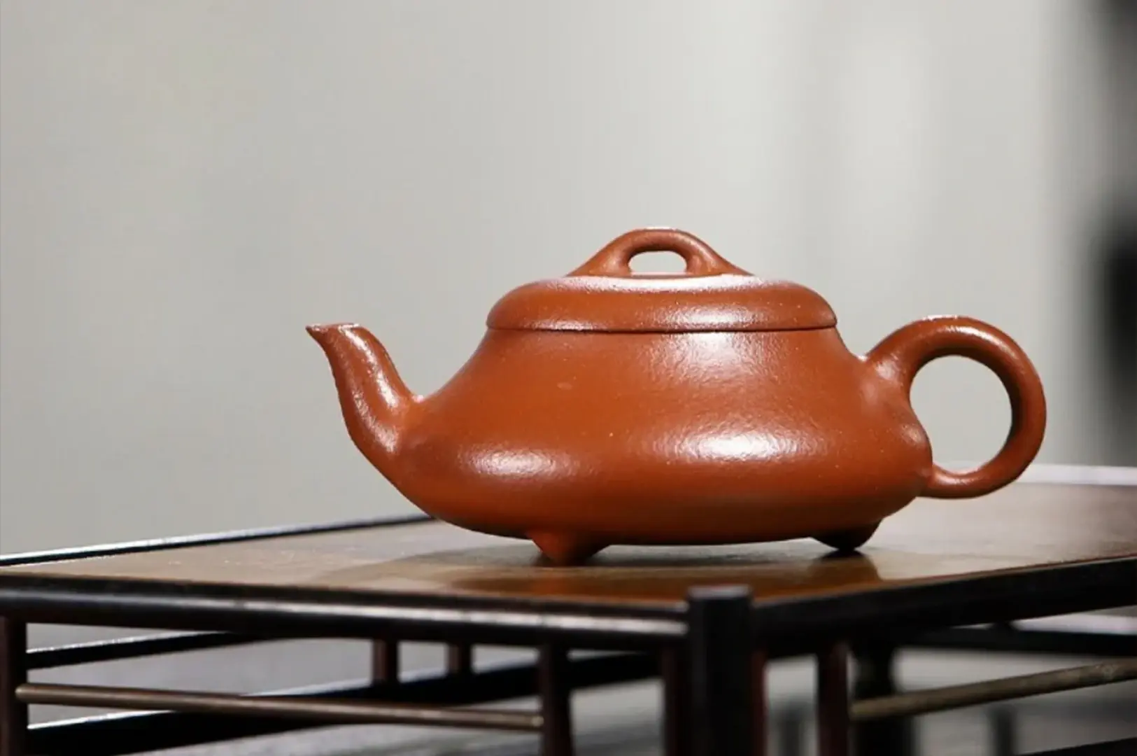 A side-profile studio shot of a rustic, earthy brown clay teapot sitting on a wooden frame. The teapot has a simple, rounded design with a short spout and a handle, resting on three small feet. The surface is smooth and matte, highlighted by a subtle sheen from the lighting.