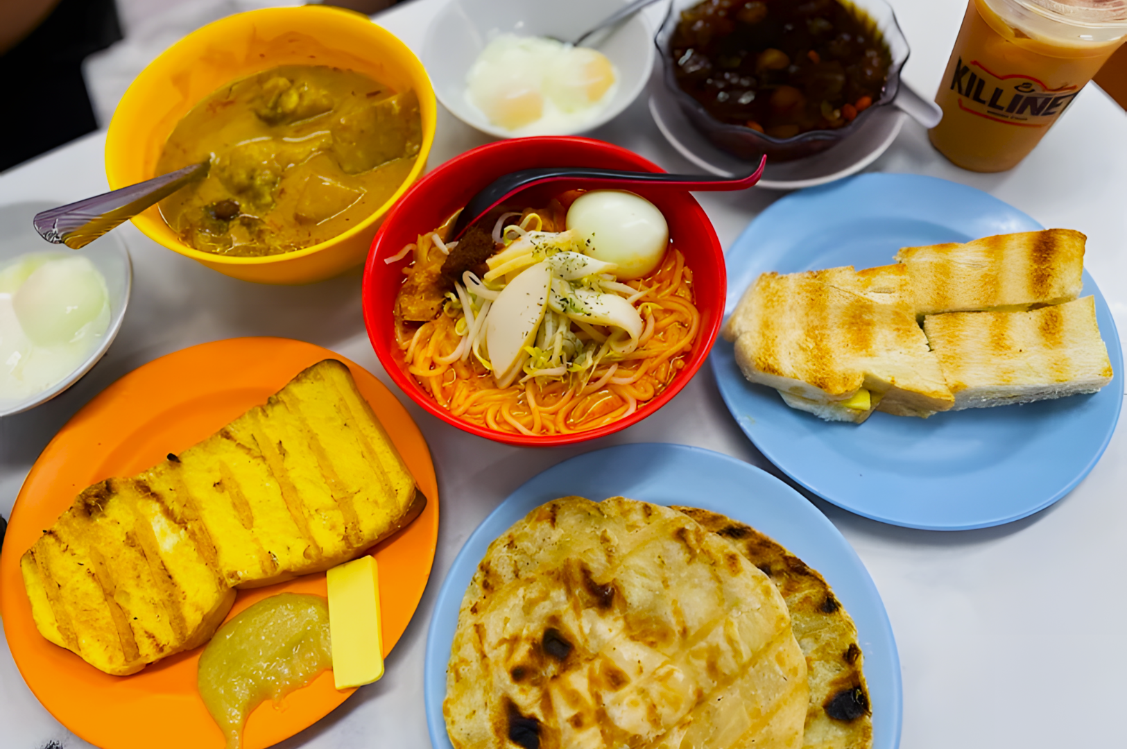 A colorful spread of Singaporean breakfast dishes on a table includes laksa with eggs, kaya toast, curry with bread, and iced tea, conveying a joyful, appetizing vibe.