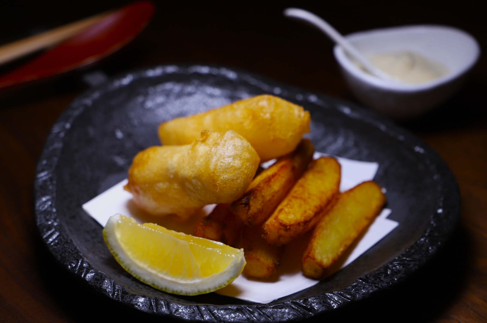 A black plate with tempura-battered fish, chunky fries, and a lemon wedge on a paper liner. A bowl of creamy dip is blurred in the background.