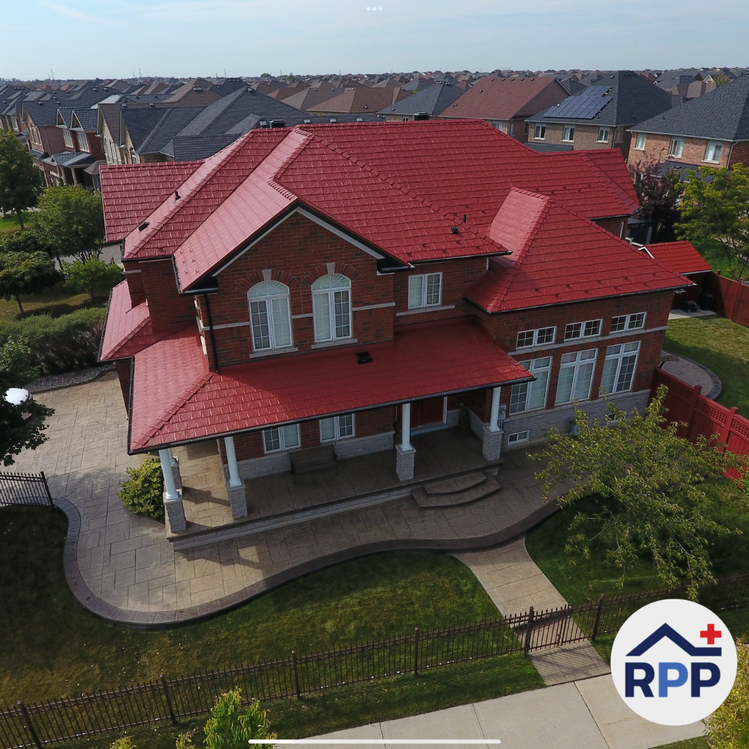 Aerial view of a red, metal-roofed, two-story brick house with white windows and pillars. It's set in a suburban neighborhood with similar homes. The Roof Pros Plus logo sits in the bottom right corner of the graphic.