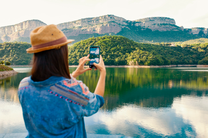 Young woman with dark hair wearing a straw hat taking a photo of a lake and mountains. 
