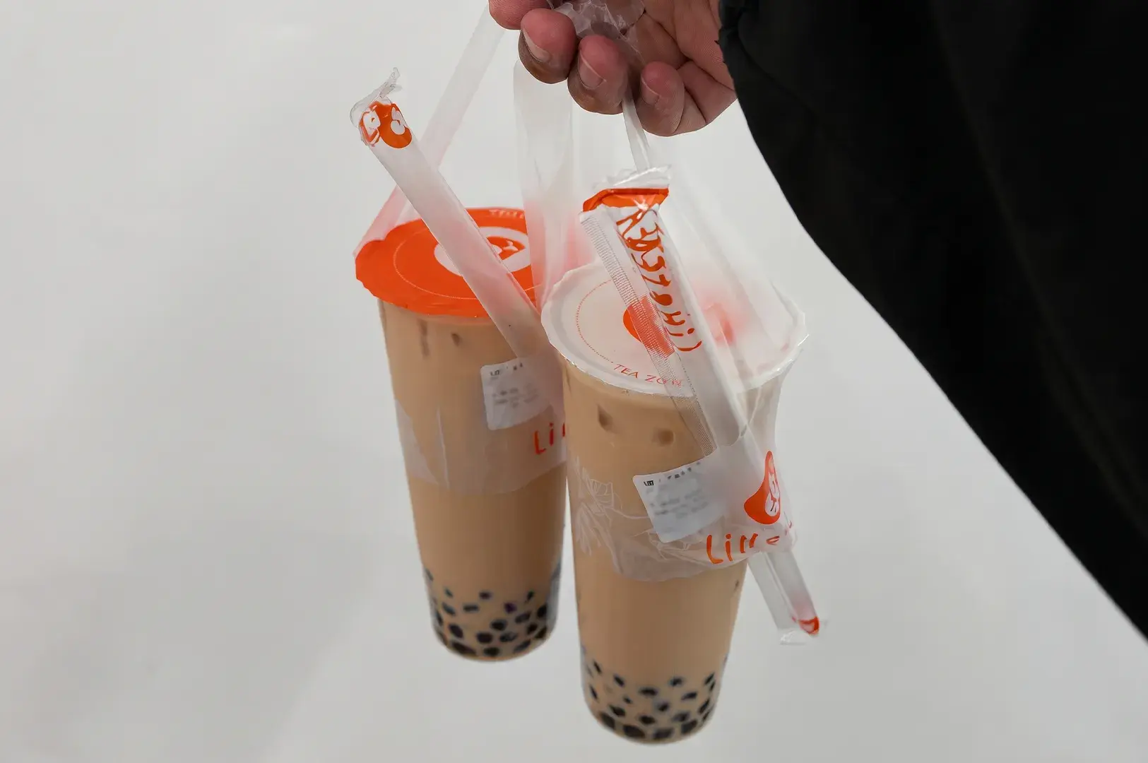 Hand holding two clear plastic takeaway bags containing LiHO bubble milk teas with tapioca boba pearls and sealed lids with straws attached.