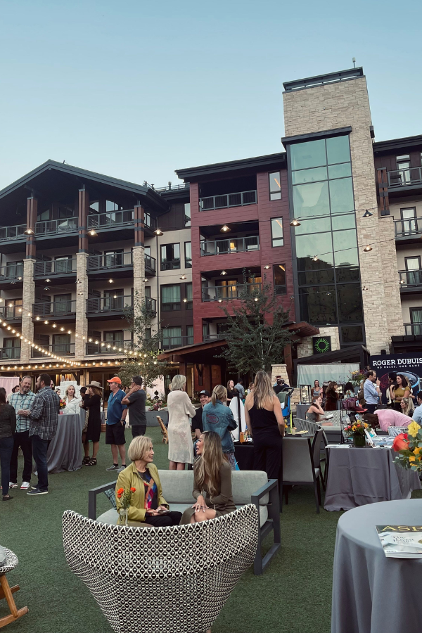 Large outdoor networking event with people mingling at cocktail tables in front of a modern hotel building, featuring string lights and evening ambiance