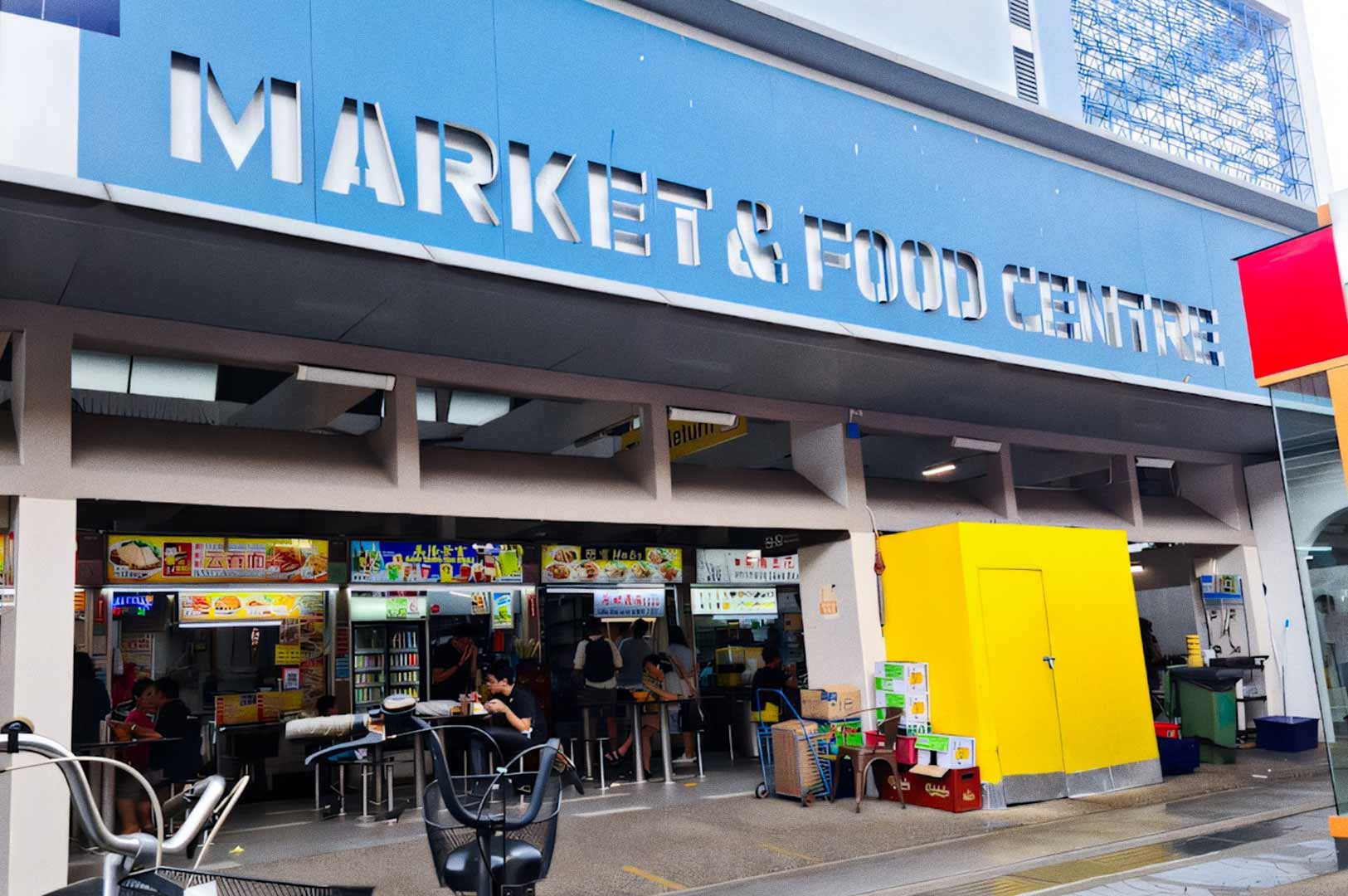 Front view of Marine Parade Market & Food Centre, showing its blue façade with large white lettering. People sit at hawker stalls inside, with colorful signboards and a bright yellow structure near the entrance.