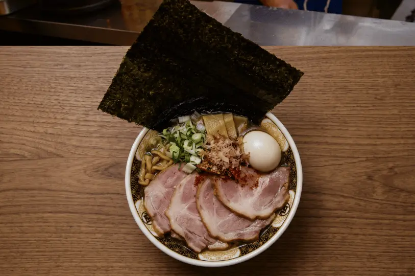 An overhead view of a steaming bowl of Kamukura ramen on a dark slate surface, featuring black chopsticks lifting a bundle of straight noodles from a golden broth packed with sweet shredded cabbage and topped with a large slice of tender pork.