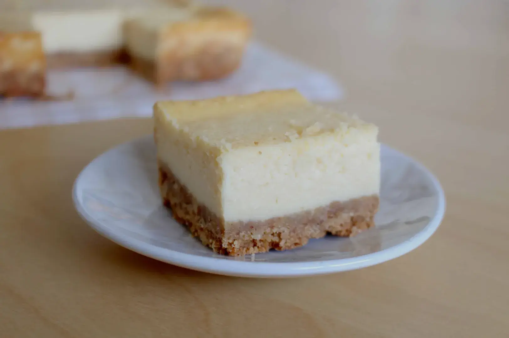 A slice of creamy cheesecake with a golden-brown crust sits on a white plate. The background shows more cheesecake pieces on a wooden table.