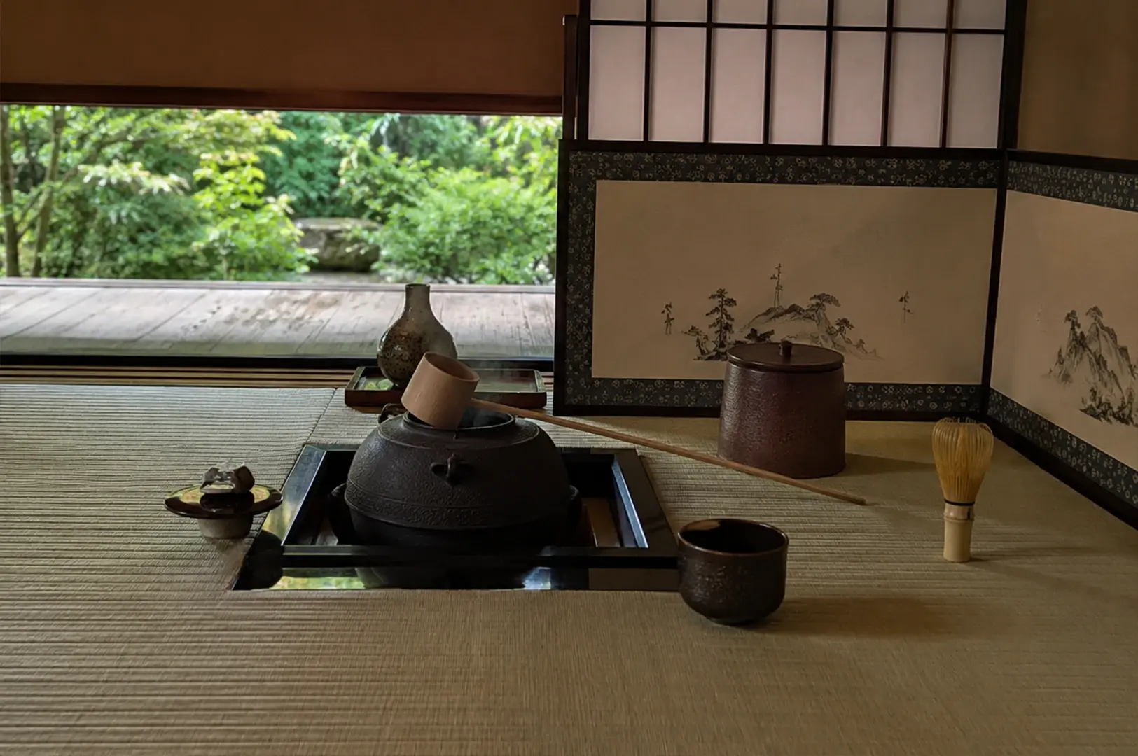 A serene indoor view of a traditional Japanese tea ceremony room featuring tatami mat flooring. In the center, a dark, heavy iron kettle rests inside a square, sunken hearth with a wooden ladle balanced across its opening. Surrounding the hearth are essential tea utensils, including a dark ceramic tea bowl, a standing bamboo whisk, and a covered container. In the background, a beautiful folding screen adorned with a minimalist ink painting of mountains sits in front of a wide-open window that reveals a lush, vibrant green garden outside.