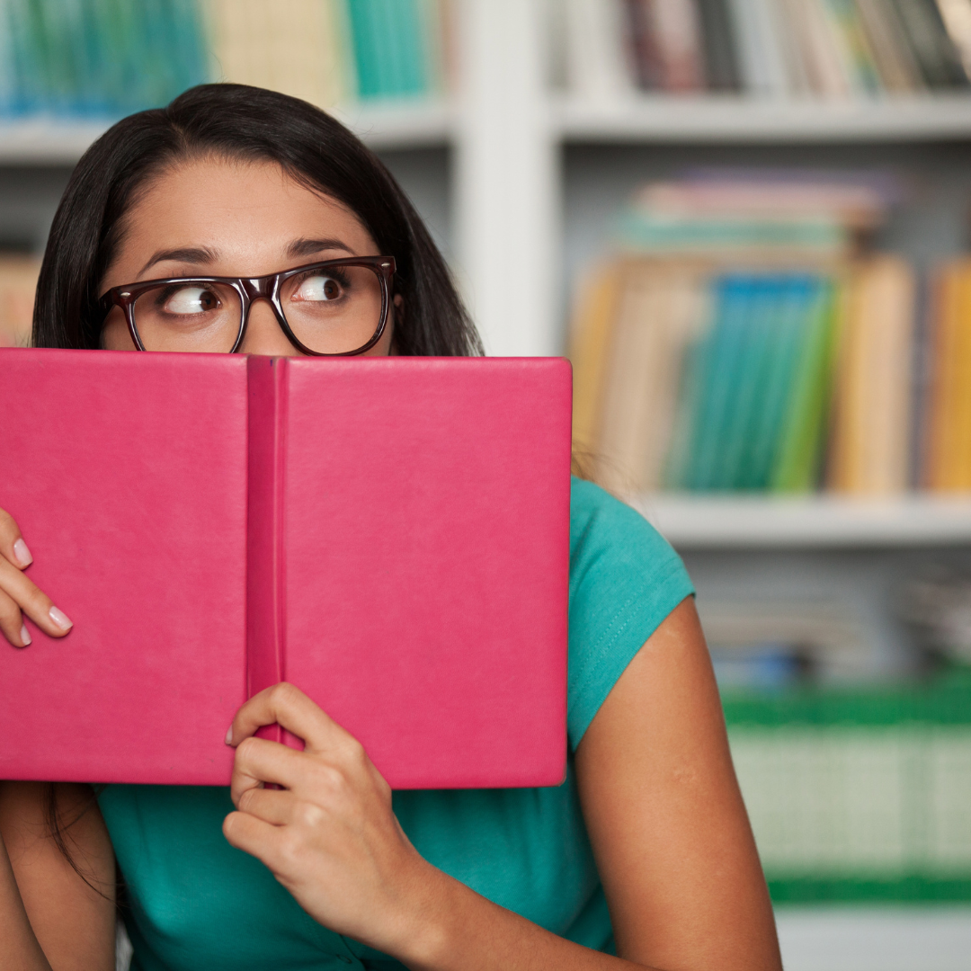 A young woman in glasses hides behind a pink book, peeking out and looking anxious.