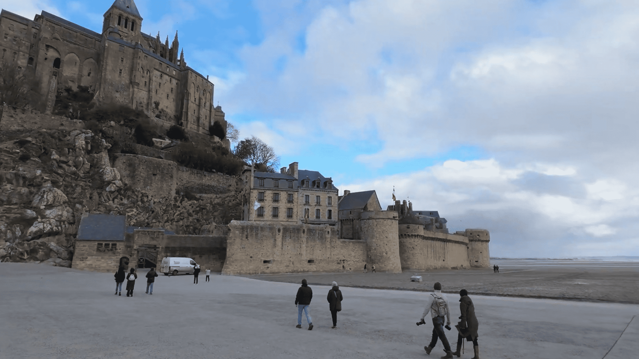 Besucher laufen über das Watt zur Burg, während die beeindruckende Abtei im Hintergrund auf einem Klosterberg thront. Die Schönheit der Landschaft und der Blick auf das Wattenmeer schaffen eine einzigartige Atmosphäre, die die Geschichte und das Welterbe dieser Region widerspiegelt.