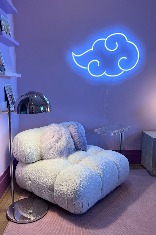 Soft lavender wall with a glowing neon cloud light above a plush boucle lounge chair, styled with a chrome floor lamp, acrylic side table, and pale pink rug.