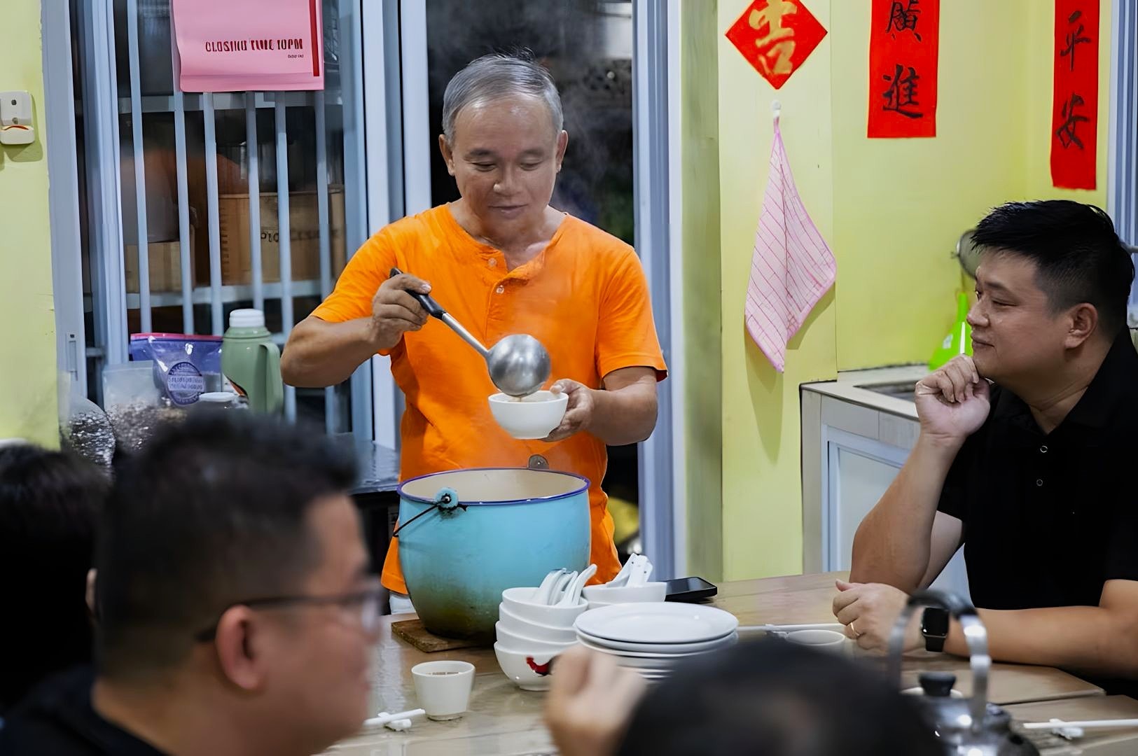 A man in an orange shirt serves soup from a large pot into a bowl in a casual dining setting. Others sit around, watching with anticipation. Walls have red Chinese decor.