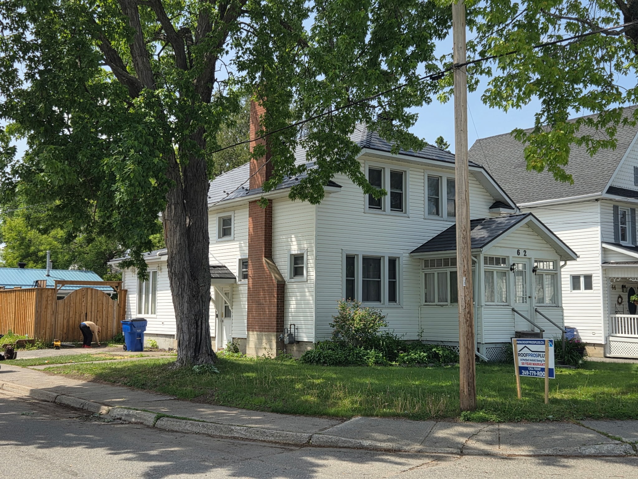 A white, two-story house with a brick chimney and metal roof is in the image, surrounded by trees. A "Roof Pros Plus" sign sits in the lawn at the front of the house.