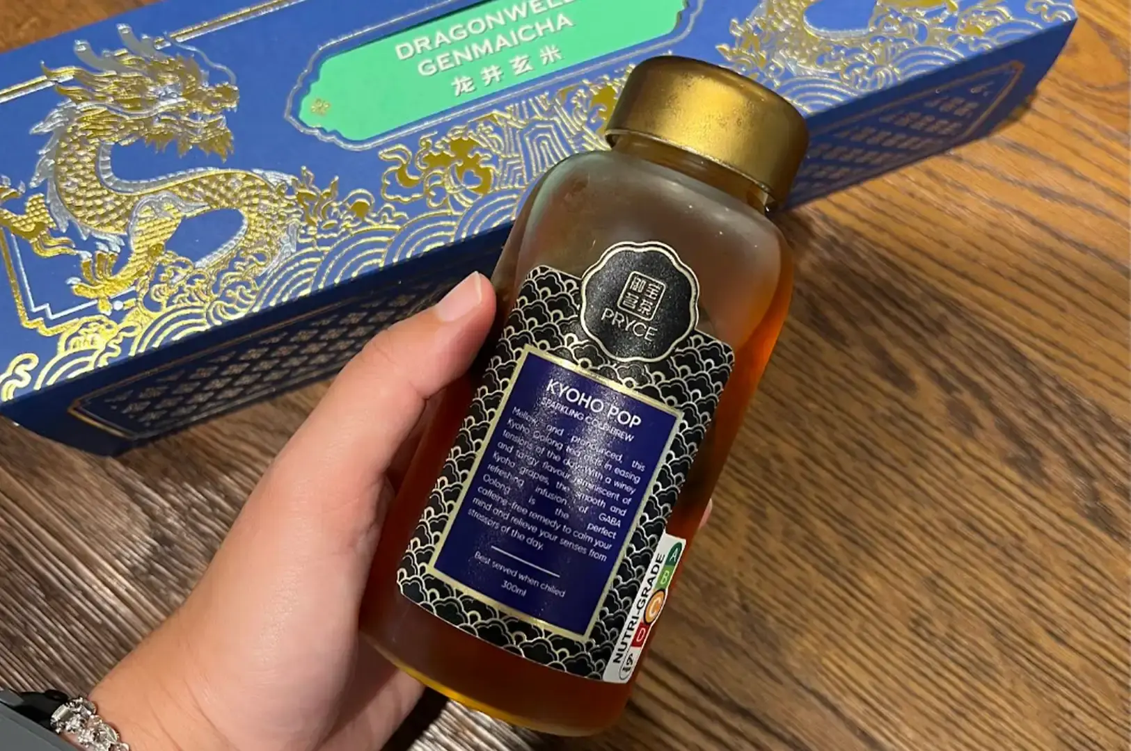 A hand holds a clear glass bottle of "KYO HO POP" sparkling cold brew tea with a gold cap and a navy blue label against a wooden background. In the backdrop, a bright blue Pryce gift box decorated with ornate gold dragon illustrations rests horizontally on the table.