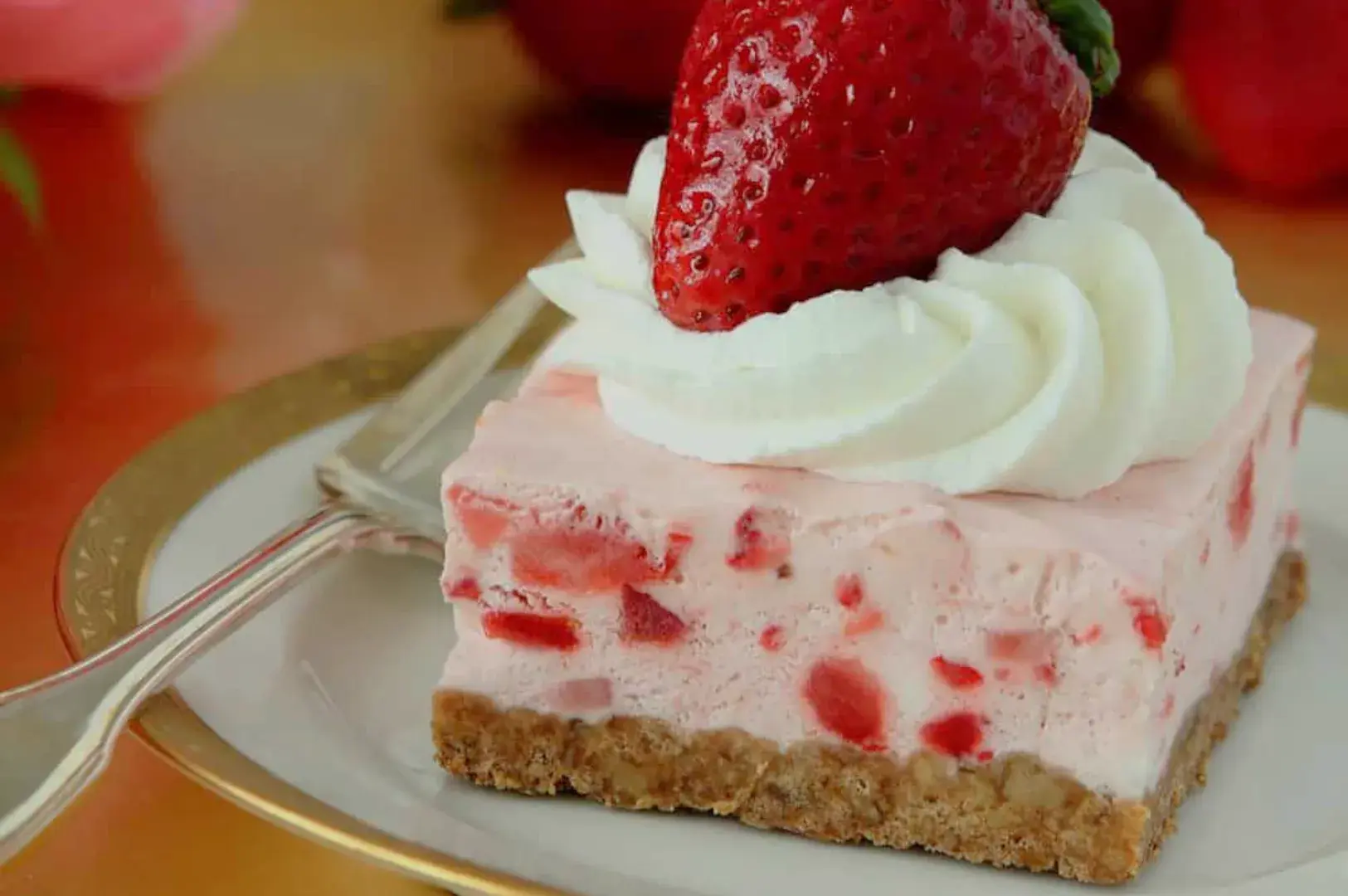 A slice of strawberry cheesecake topped with whipped cream and a fresh strawberry, served on a white plate with a gold rim.