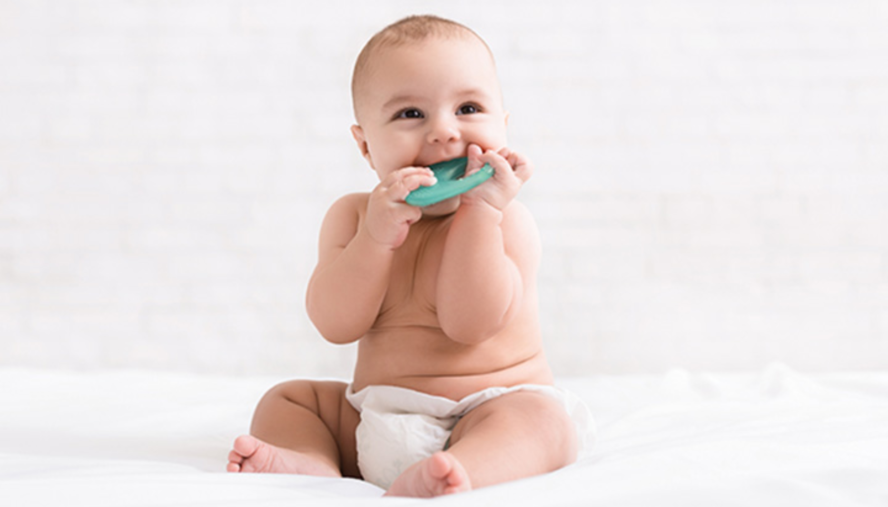 Baby sitter på hvitt teppe med bleie og tygger på grønn bitering for tannfrembrudd.