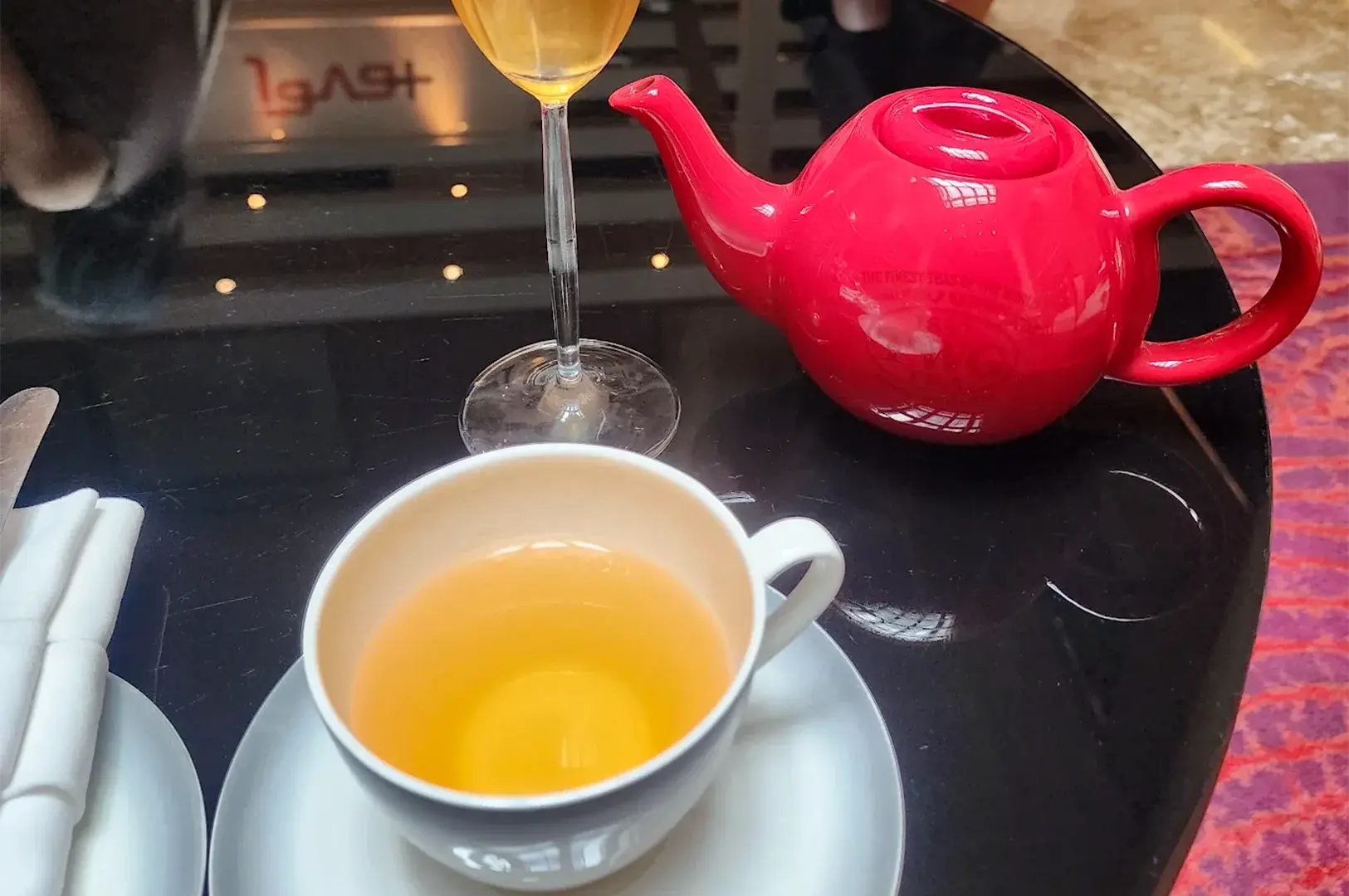 A vibrant red TWG teapot alongside a teacup containing clear amber tea and a long-stemmed glass on a glossy black lounge table.