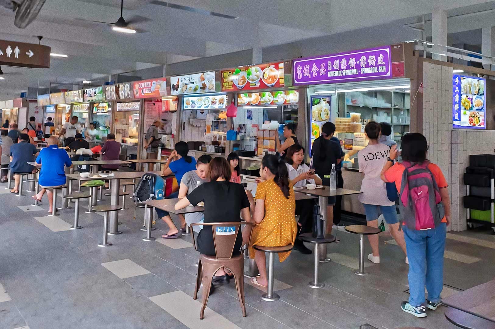 Indoor hawker centre with rows of food stalls displaying colorful signboards. Diners sit at metal tables and stools while others queue at various stalls. The space is bright and bustling with activity.