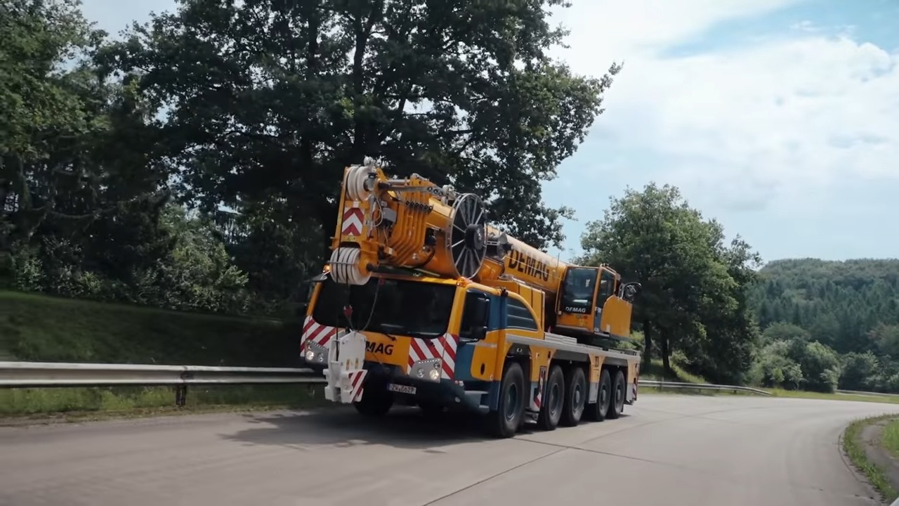 Large yellow and blue Demag mobile crane traveling on a scenic road surrounded by lush green trees