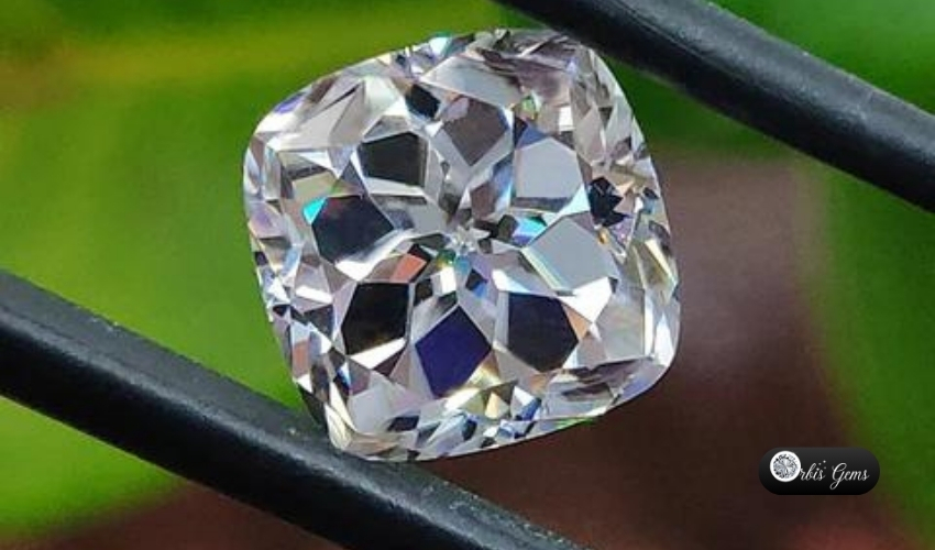 Cushion-cut moissanite gemstone held by tweezers, showcasing brilliant facets and fire, with a natural green backdrop and Orbis Gems logo.