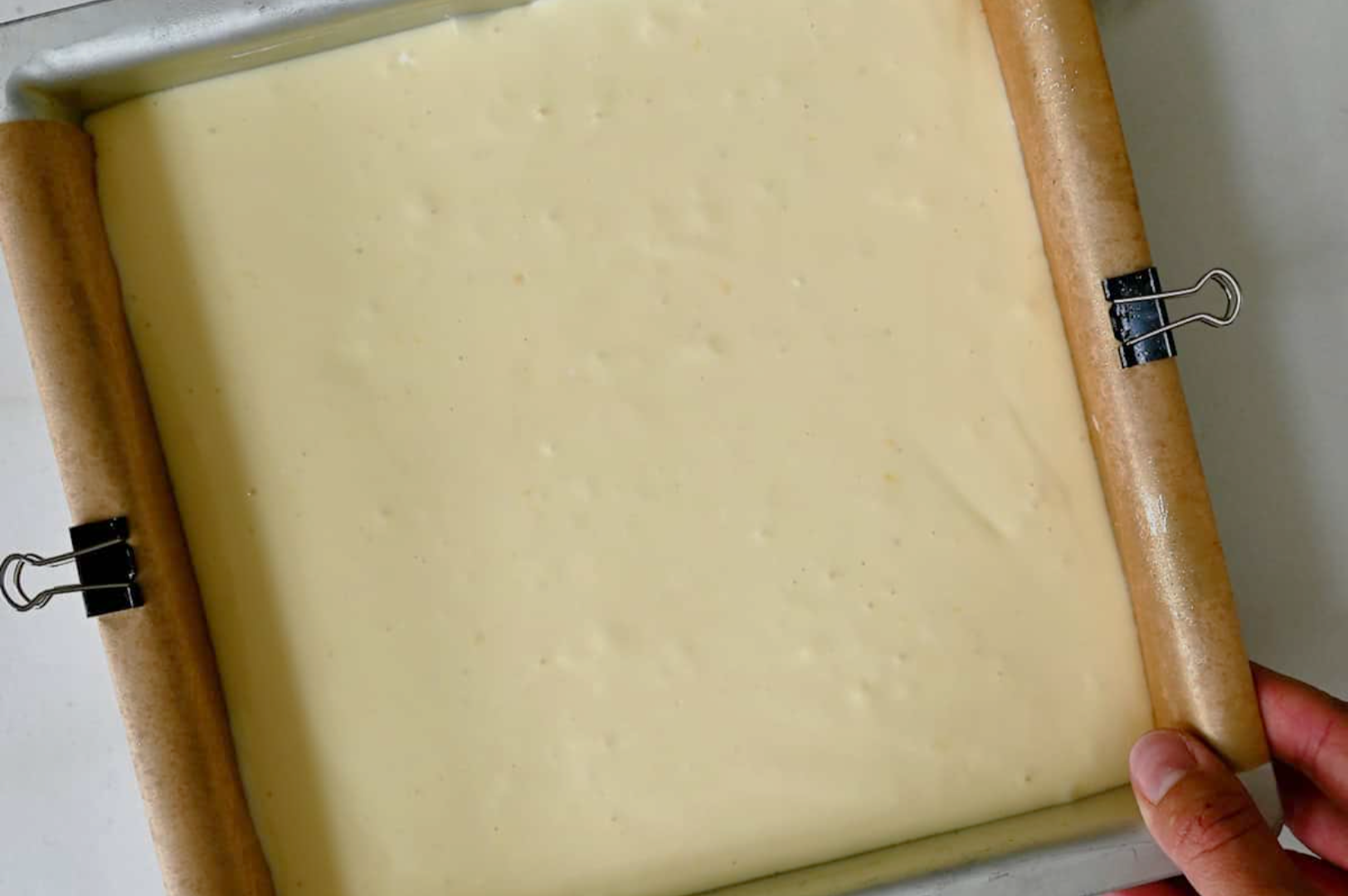 Close-up of a square baking pan lined with parchment paper, secured with clips, filled with pale, creamy batter. A hand holds the pan, ready for baking.