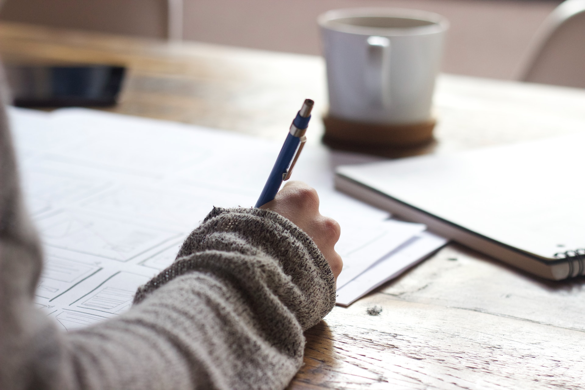 person writing on a book on a wooden table with ceramic mug