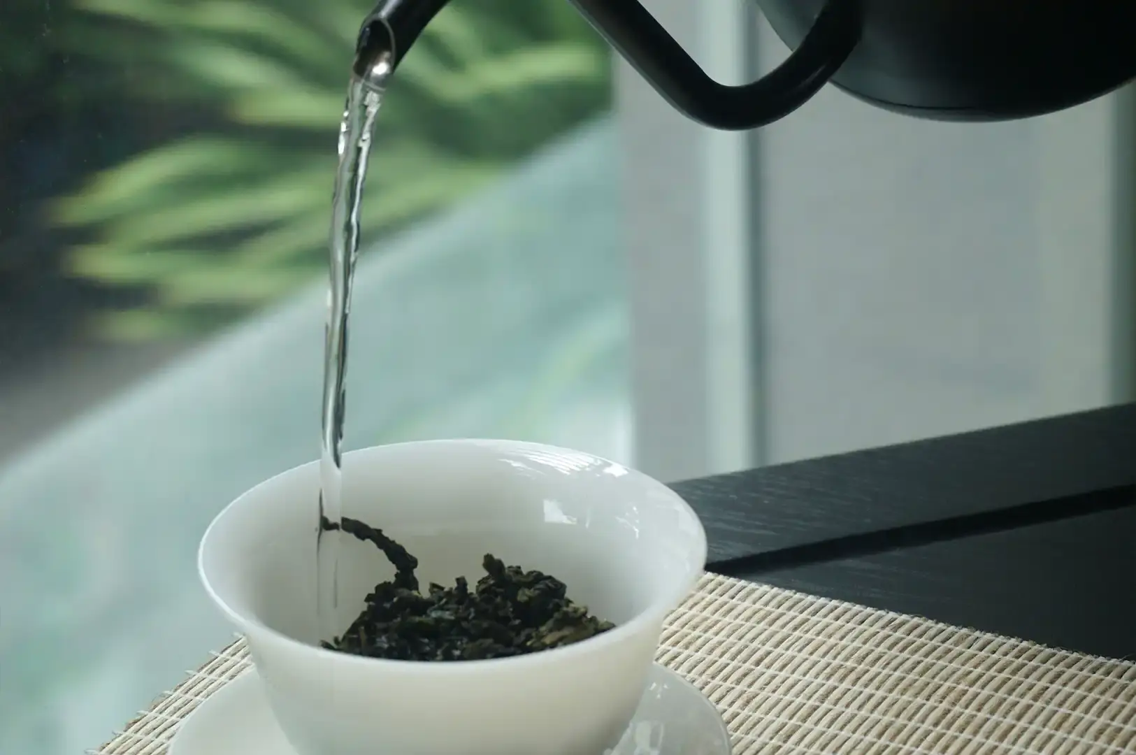 A black tea kettle pours a precise, thin stream of hot water into a white tea bowl resting on a woven bamboo mat. The bowl is partially filled with dark, curled Tie Guan Yin tea leaves, which begin to gently unfold as they come into contact with the hot water, set against a blurred, serene outdoor background.