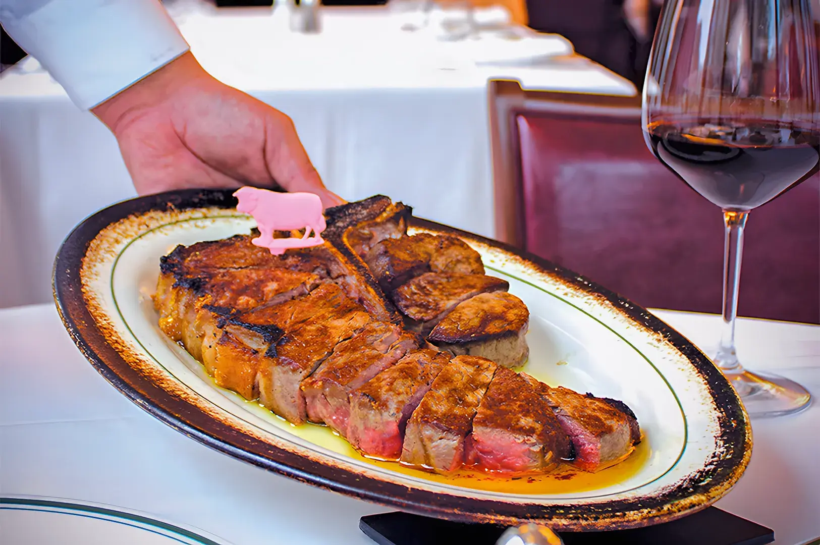 A hand serves a juicy, grilled steak on a white platter with a small pink cow figurine. A glass of red wine sits nearby on a white tablecloth.