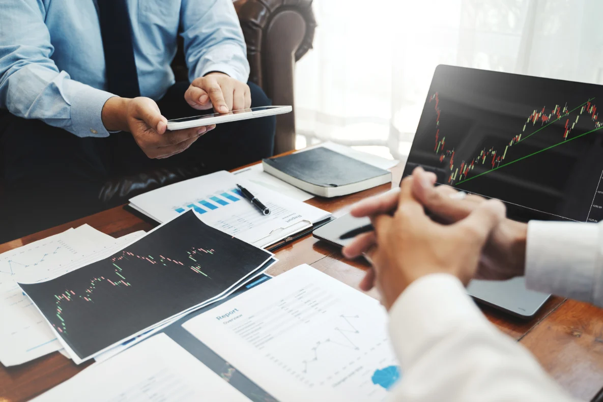 Two people are reviewing financial charts and data on a table, with one person holding a tablet and the other looking at a laptop screen displaying stock market trends.
