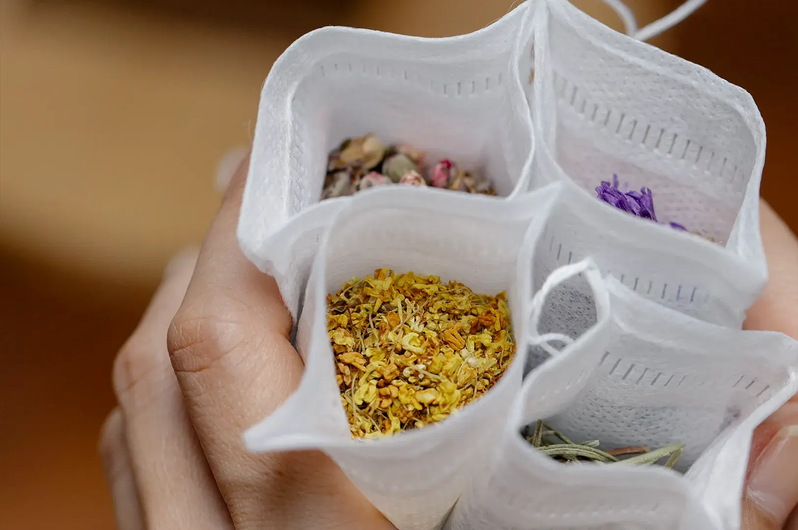 A close-up shot focusing on a person's hand holding open a multi-compartment white mesh pouch, revealing various colorful dried botanicals inside each section. The compartment closest to the camera is filled with vibrant yellow, finely ground osmanthus or chamomile flowers, while other visible compartments contain pink rosebuds, purple petals, and green herbal leaves. The background is softly blurred, keeping the emphasis on the textures and colors of the herbal ingredients.