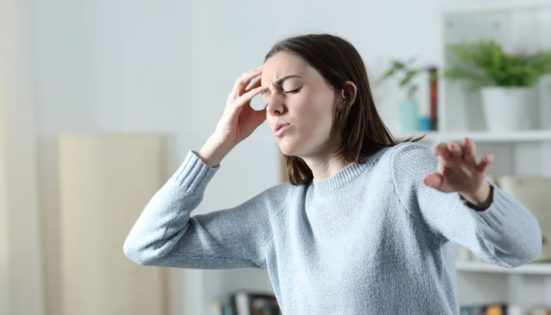 Kvinne med lukkede øyne holder hodet med én hånd og strekker den andre ut, tydelig preget av svimmelhet eller hodepine.
