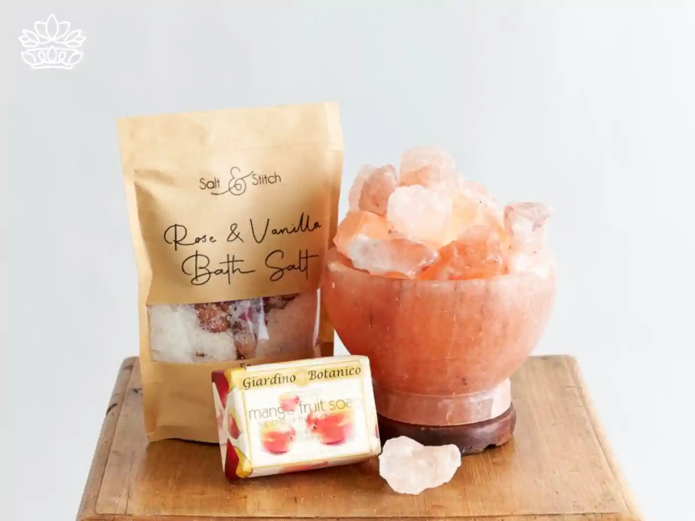 A soothing display of wellness products including Rose & Vanilla bath salt in a paper pouch and a mango fruit soap beside a salt lamp on a wooden surface, part of the mental health gift boxes by Fabulous Flowers and Gifts. Delivered with a focus on nurturing and relaxation.