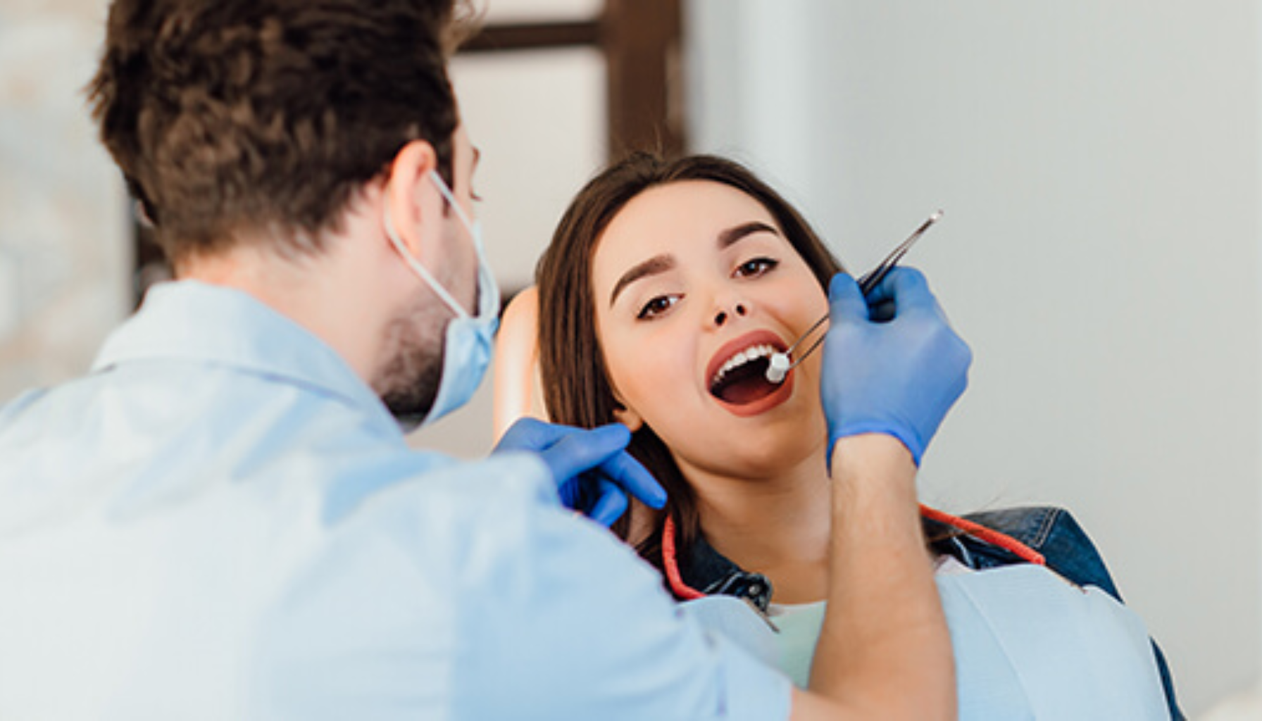 Tannlege med munnbind og blå hansker undersøker åpen munn hos kvinnelig pasient i tannlegestol i lyst behandlingsrom.